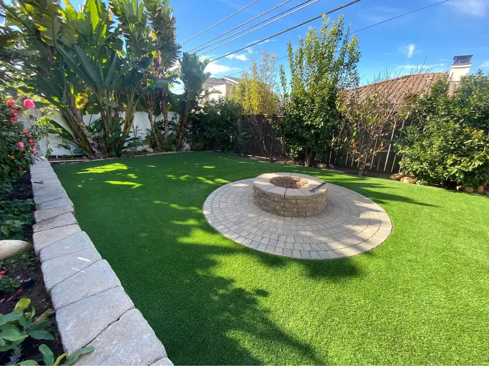 A backyard with artificial grass installed by a Turf Installer Arcadia AZ, featuring a circular stone fire pit on a patio, surrounded by lush plants, trees, and a wooden fence under a clear blue sky.