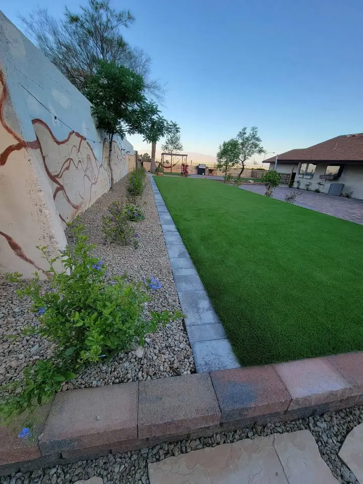 A backyard with a green artificial lawn by a Turf Installer Phoenix AZ, bordered by a stone path and gravel with small flowering plants. A mural brightens the left wall, while the house is visible to the right.