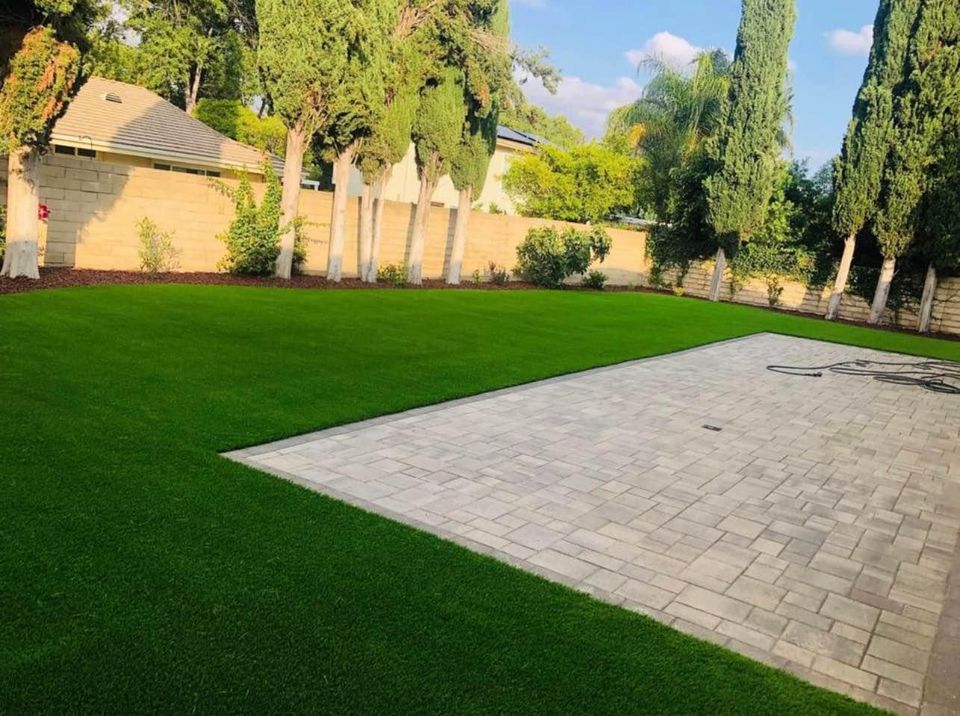A backyard with a neatly trimmed green lawn, a large paved patio area, and tall trees along the edges, bordered by a tan brick wall under a sunny sky.