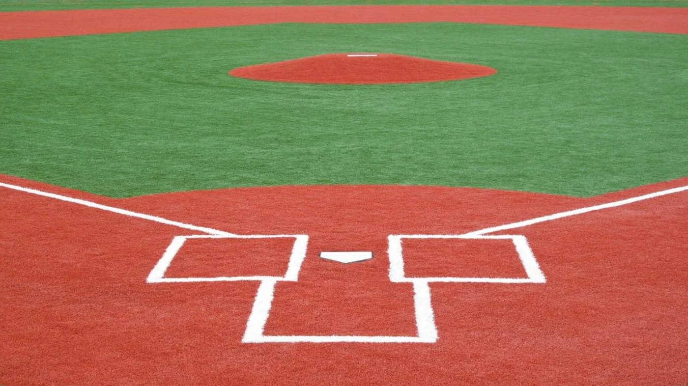 A baseball field’s home plate area and pitcher’s mound are shown, with red artificial turf and white chalk lines outlining the batter’s boxes and bases. The green turf marks the infield grass.