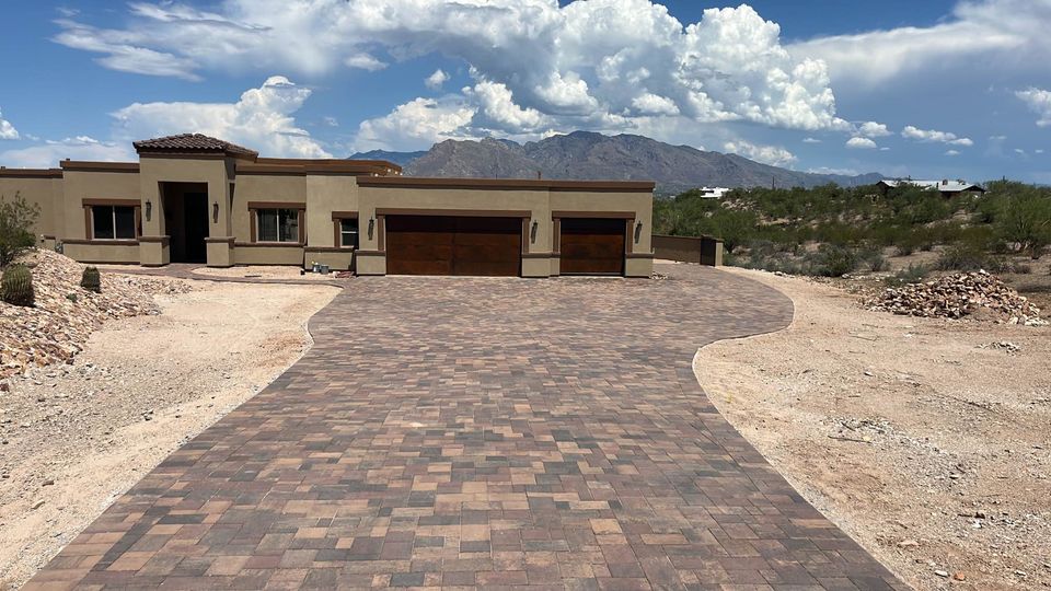 A modern single-story house with a wide, curved brick driveway, set in a desert landscape with sparse vegetation and mountains in the background under a partly cloudy sky.