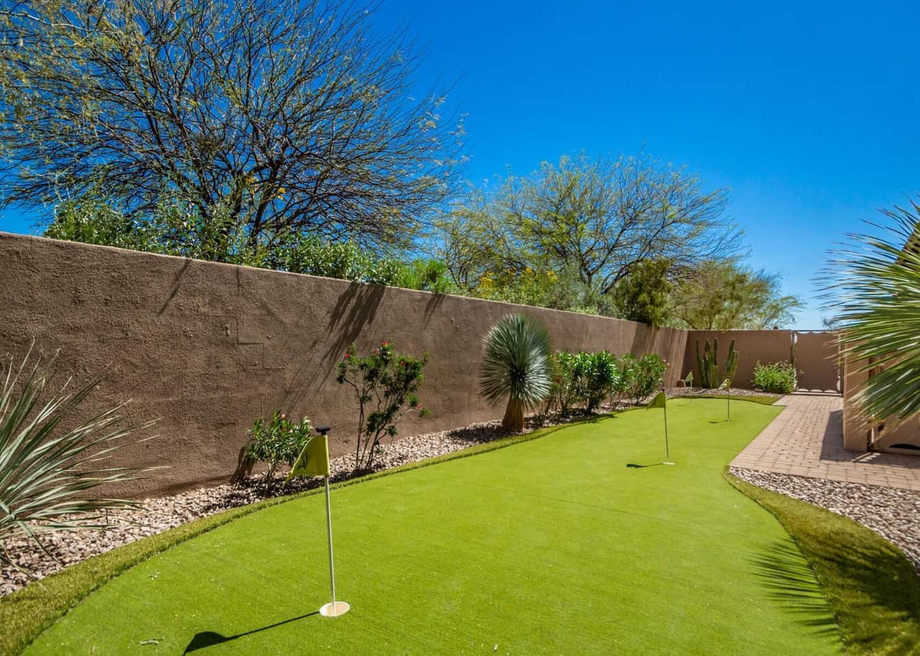 A backyard with a small putting green, three golf flags, desert plants, and trees along a stucco wall under a clear blue sky.