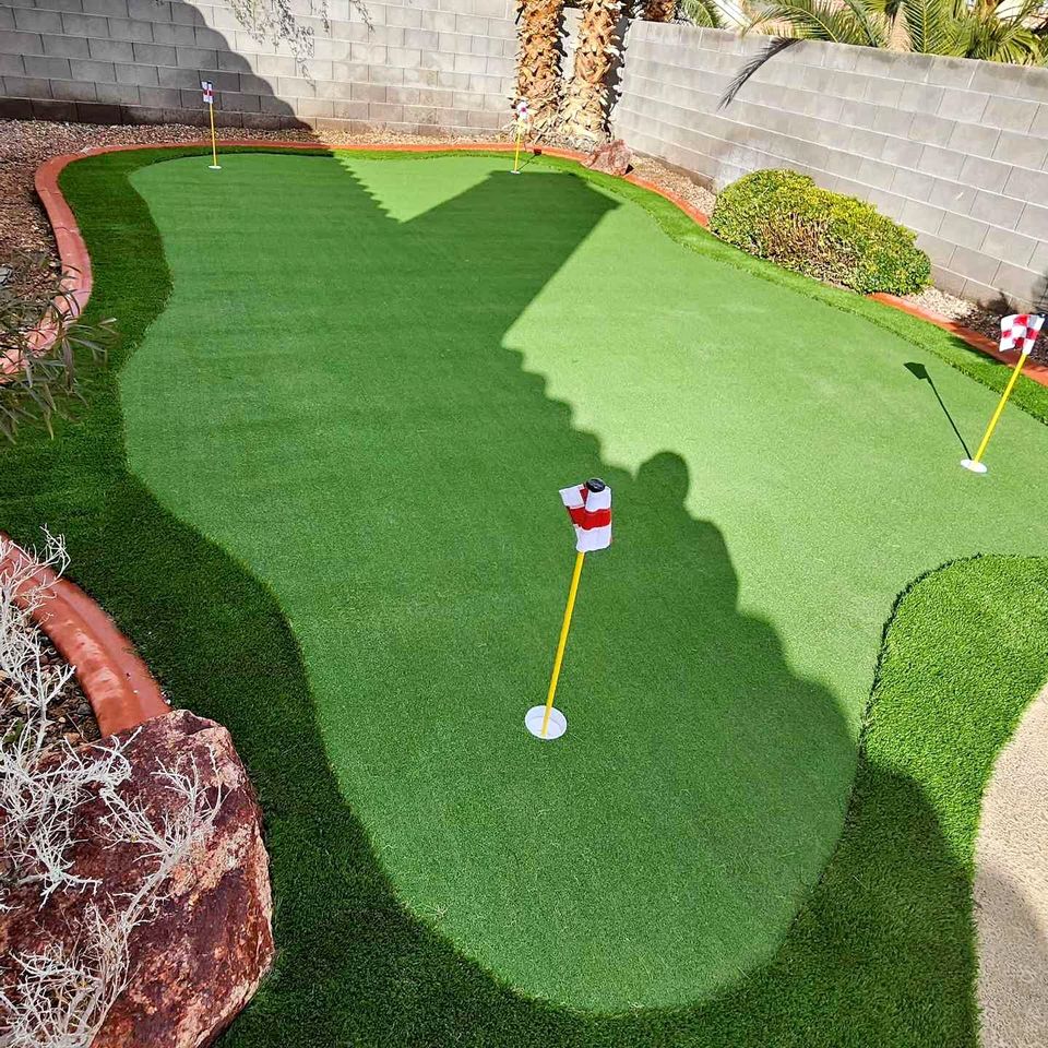 A sunny backyard putting green with artificial turf, three small flagsticks with red and white flags, surrounded by decorative edging, plants, and a block wall. A shadow of a palm tree falls across the green.