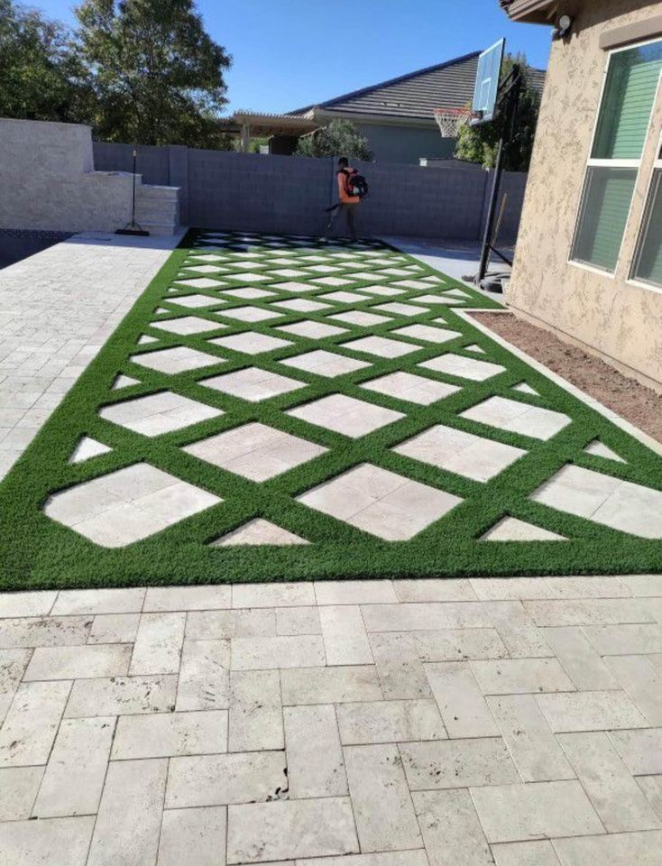 A backyard features a patio with a diamond-shaped grid pattern of concrete pavers and artificial grass, bordered by a wall and a house. A person in the background uses a leaf blower.