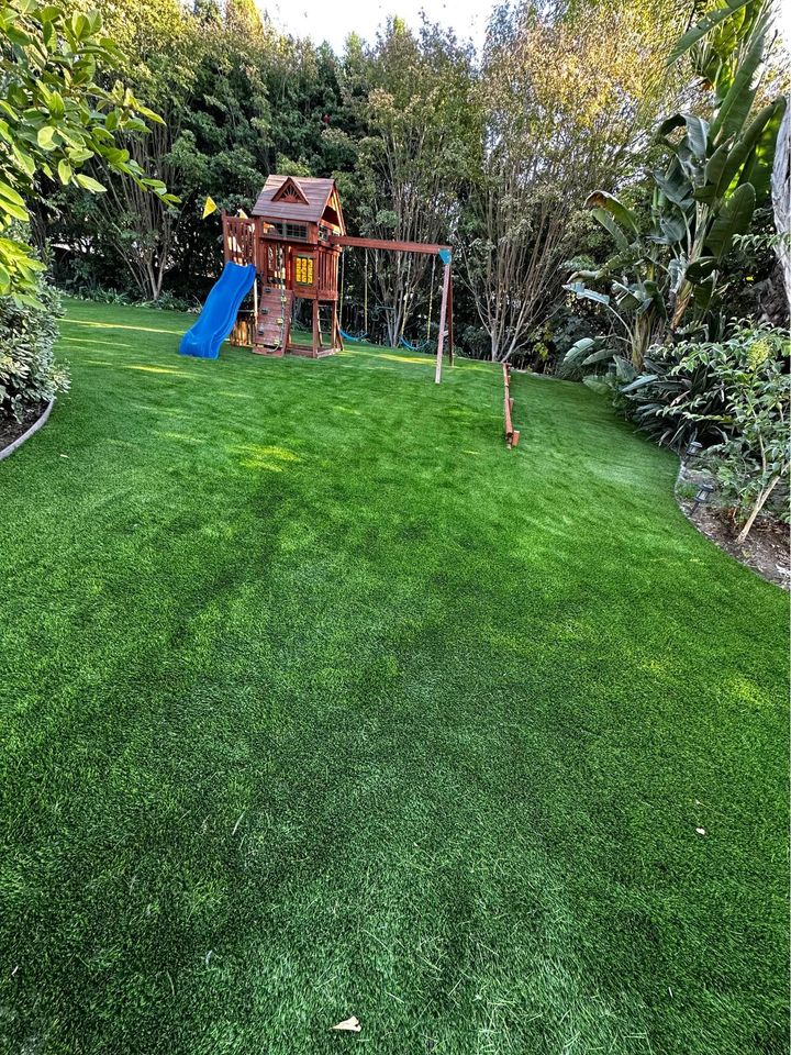 A backyard with lush green grass features a wooden playset that includes a slide and swings, surrounded by dense trees and bushes.