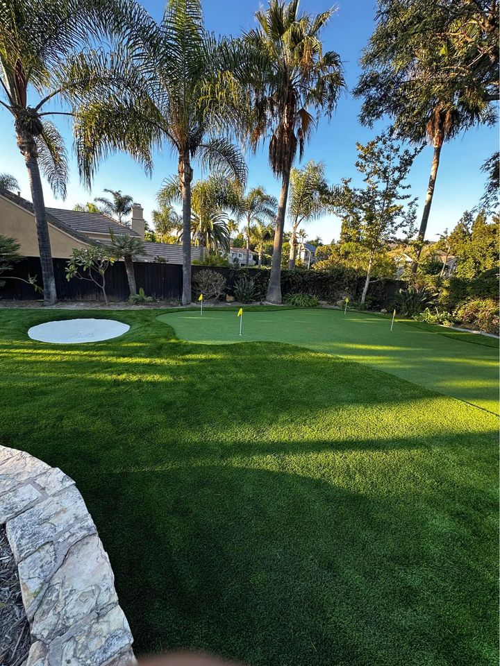 A backyard putting green with several yellow-flagged holes, surrounded by lush artificial grass, palm trees, and landscaped greenery under a clear blue sky. A stone-edged sand bunker is visible on the left.