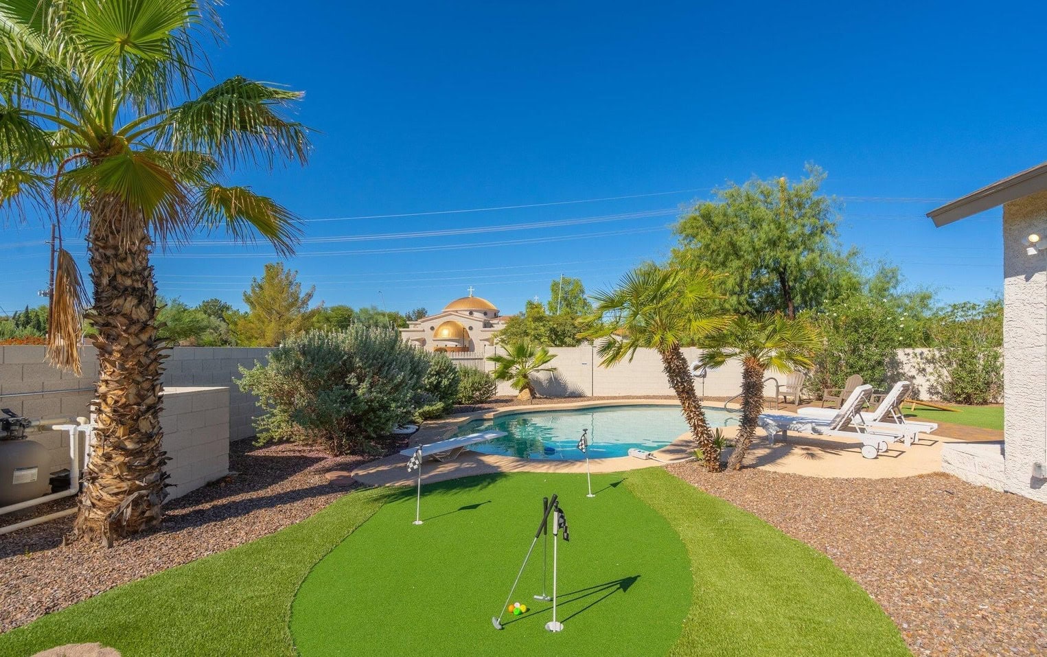 Backyard with artificial putting green, palm trees, desert landscaping, and a swimming pool with lounge chairs under clear blue sky.